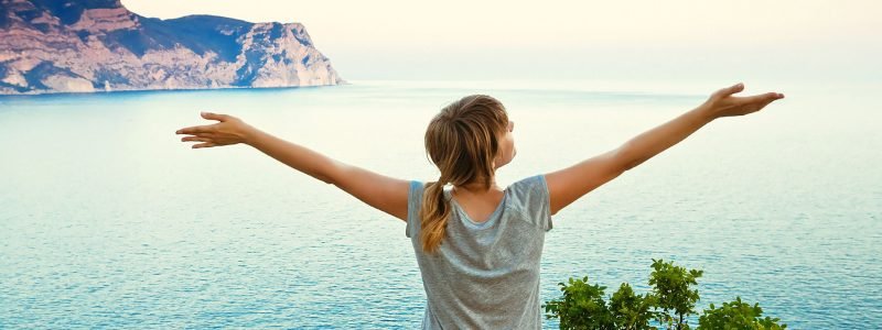girl sits on a hill with raised hands and looking at the sea